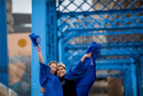  两名大学生戴着蓝色的帽子，穿着蓝色的长袍，拥抱在一起，在空中挥舞着他们的手臂. 