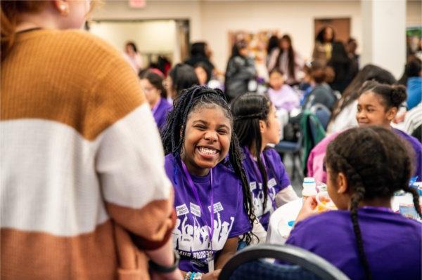 一名身穿紫色衬衫的学生在满屋子的活动参与者中微笑. 