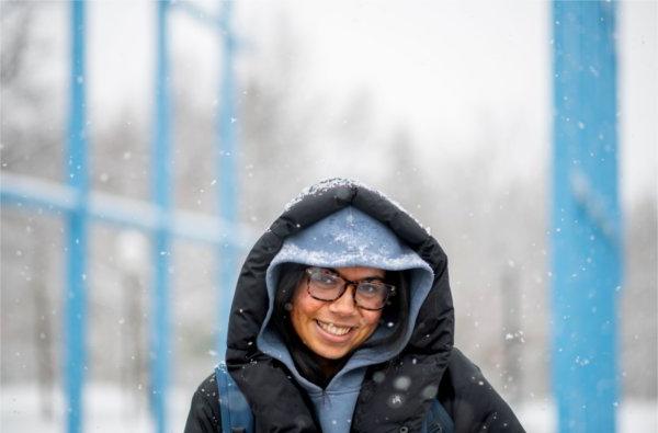 一名大学生走在大学校园里，雪花落在她蓝黑相间的兜帽和外套上，她对着镜头微笑. 