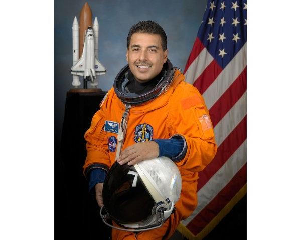 A person wearing an orange uniform and holding an astronaut's helmet smiles. An American flag and the model of a rocket are in the background.