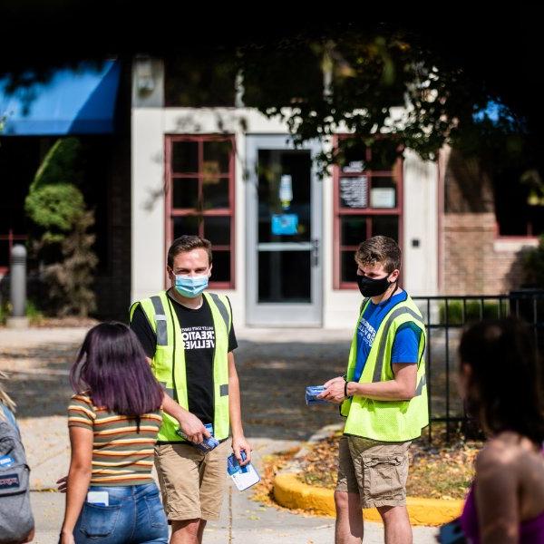 在艾伦代尔校区，两名身穿黄色背心的弗吉尼亚州立大学男生给另一名弗吉尼亚州立大学学生分发可重复使用的面罩.