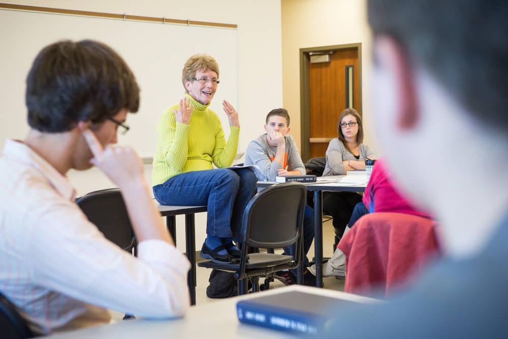 一个女人坐在课桌上，在课堂上和学生交谈