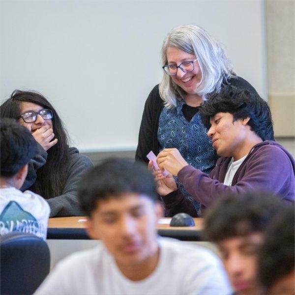 a young student shows a card to a woman leaning over his shoulder, while a classmate laughs and looks on