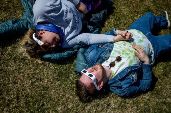 两人手牵着手躺在地上，戴着特殊的眼镜观看日食.