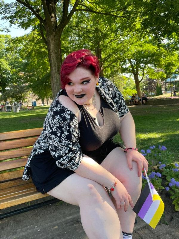 佐伊贝尔克, 长着紫红色短发的人, sits on a bench and holds a small non-binary flag with yellow, 白色, 紫色的, 黑色条纹.