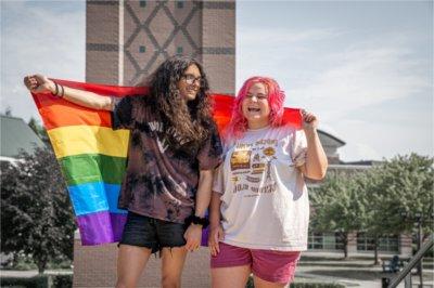两人, one with pink hair and the other with long black hair and glasses, hold a pride flag behind them and stand in front of the Cook Carillon clock tower.
