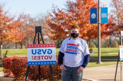 山姆·雅各布斯戴着面具站在外面GVSU投票标牌旁边穿着同样的t恤, 有GVSU的选票!