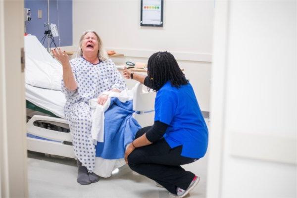 KCON学生Amarachi obh跪在“Janet”面前，这是一个标准化的 病人, 在德沃斯跨专业健康中心模拟中心的医院套房进行的试点模拟中.