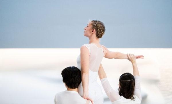 Dancers wearing while perform in front of a light blue and white wall. 